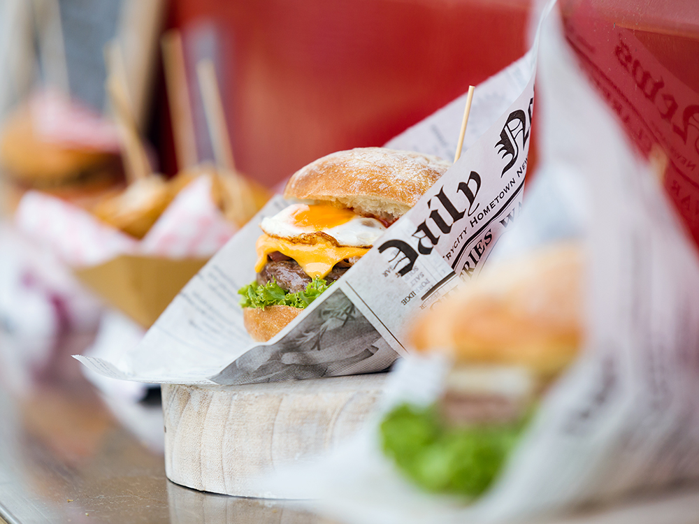 Köstliches aus dem Foodtruck: Ein Burger mit Spiegelei und zerlaufendem Käse wird in einem Burger-Papier präsentiert. Das Papier sieht aus wie eine alte Zeitung.